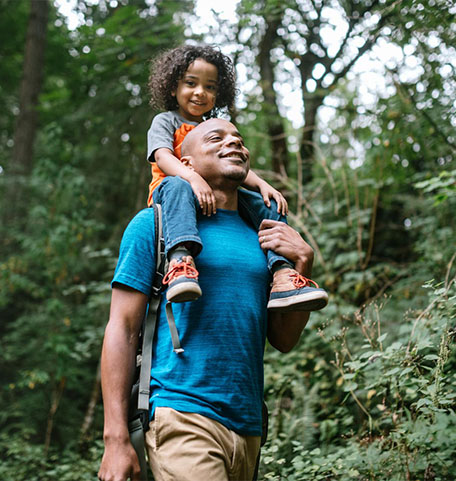 Ein Vater wandert mit seiner Tochter auf seinen Schultern durch einen Wald